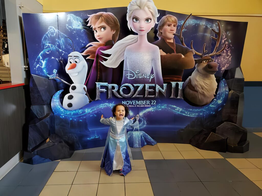 Small girl standing in front of Frozen 2 movie sign in Elsa dress